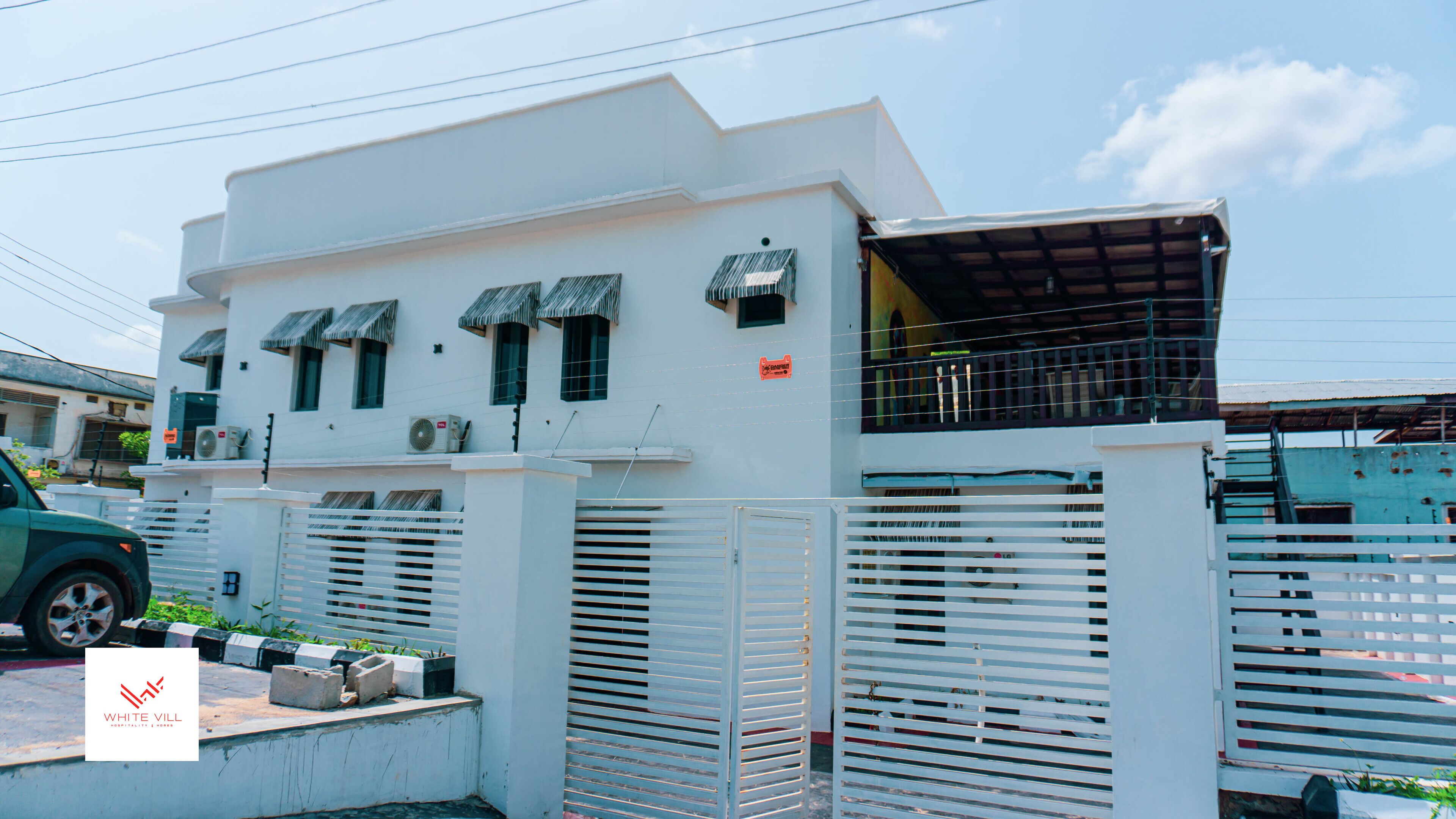 A modern white building with a gated entrance, a covered balcony, and a sign indicating a hotel.