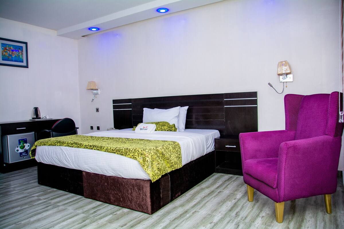 Quarto de hotel com cama, escrivaninha, cadeira e mesa de cabeceira.