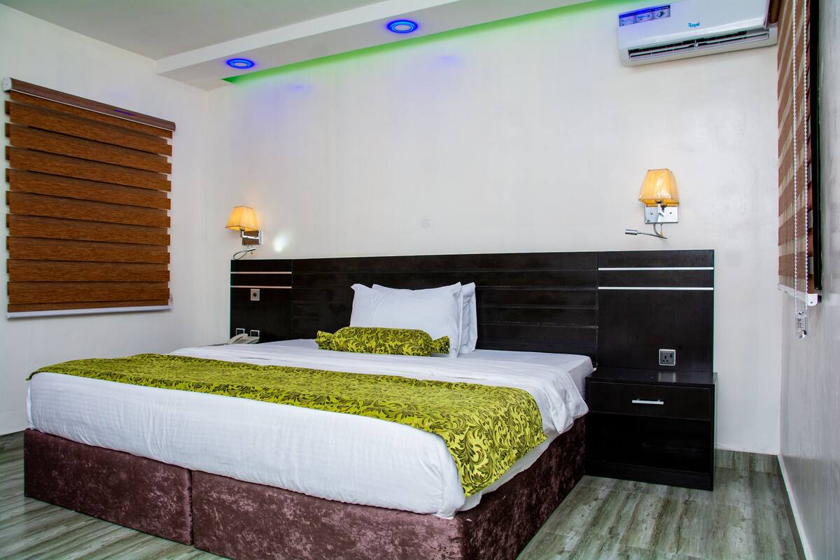 Quarto de hotel com uma cama grande, abajures, cabeceira de madeira, mesa de cabeceira e aparelho de ar condicionado.