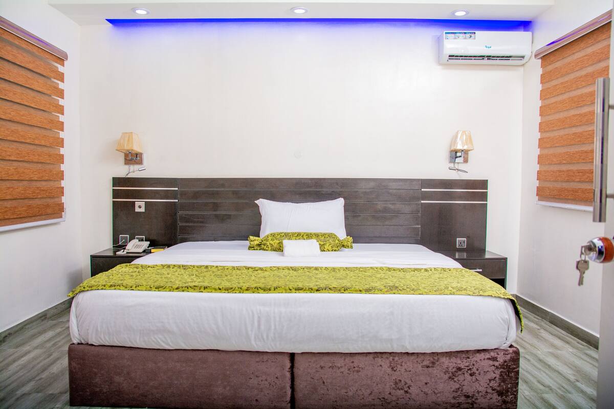 Quarto de hotel com uma cama grande, duas luminárias de cabeceira, um ar condicionado e um painel de parede de madeira.