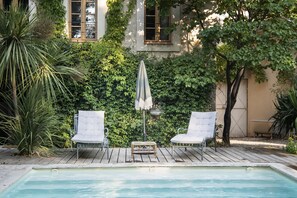 Et poolområde med to stole og en parasol, omgivet af frodig vegetation og en bygning med vinduer.