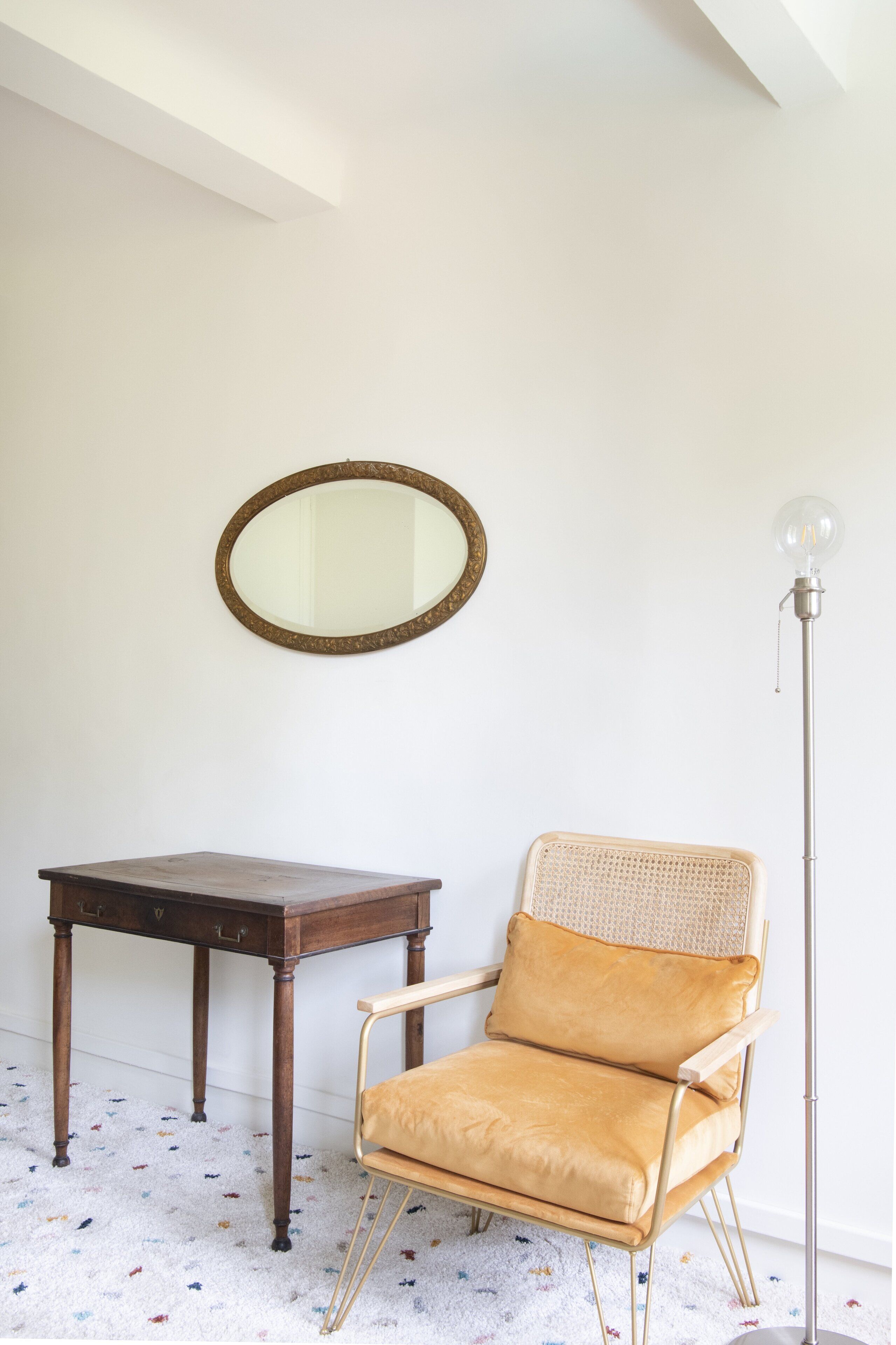 Een kamer met een houten tafel, een rieten stoel met kussen, een spiegel aan de muur en een vloerlamp.