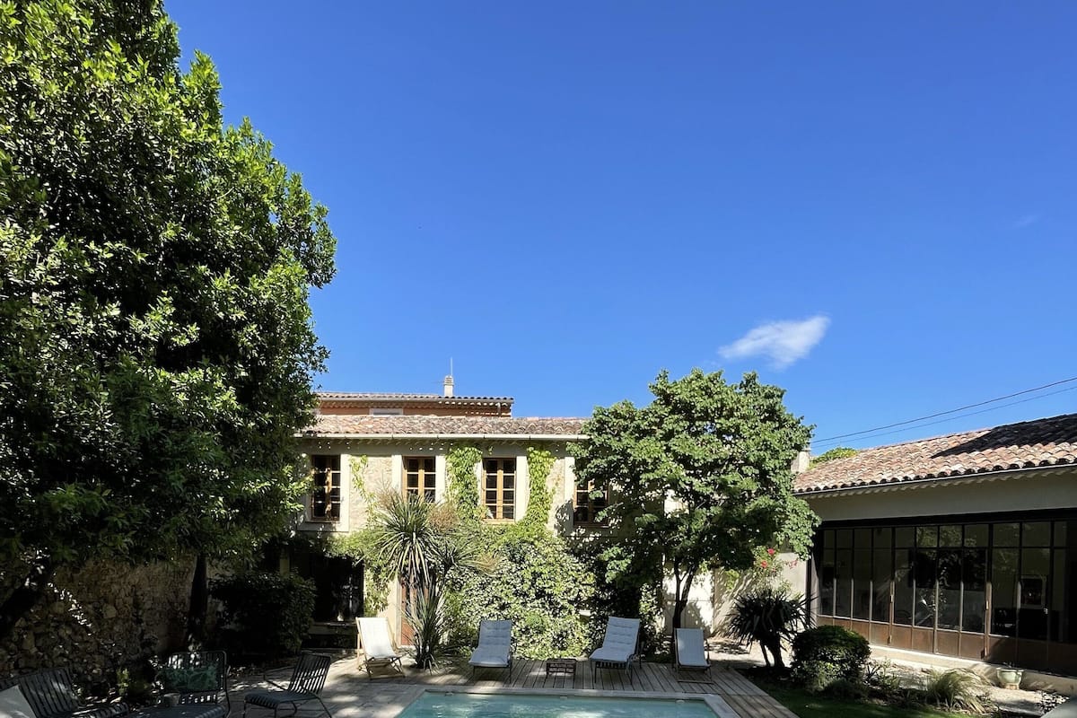 A rectangular swimming pool with a wooden deck, surrounded by lounge chairs and a stone building with a tiled roof.