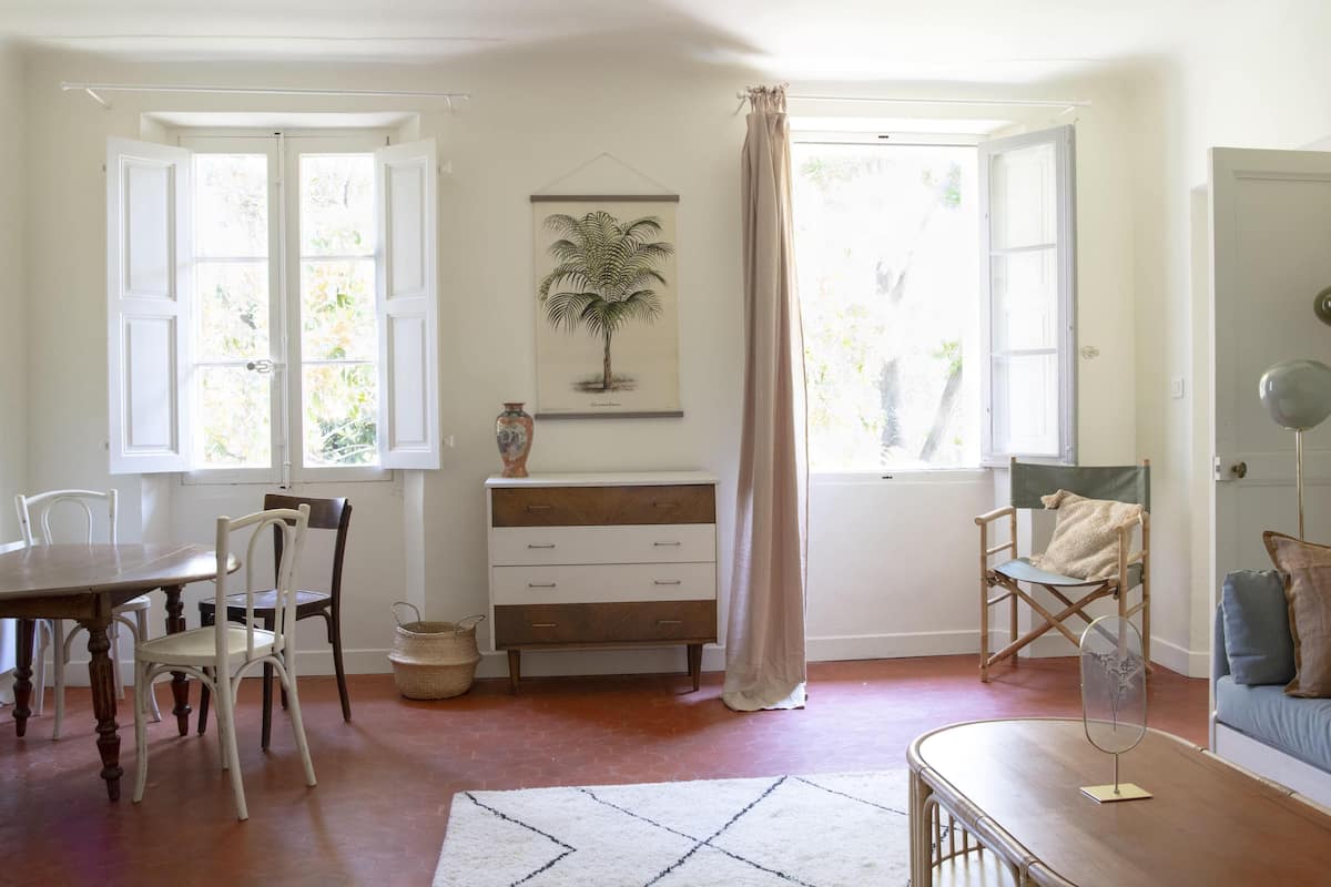 A room with a wooden dining table, chairs, a sofa, a dresser, and a large window with curtains.
