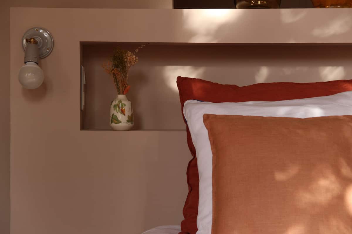 A bedroom with a bed, a wall-mounted light fixture, and a small shelf with a vase of dried flowers.