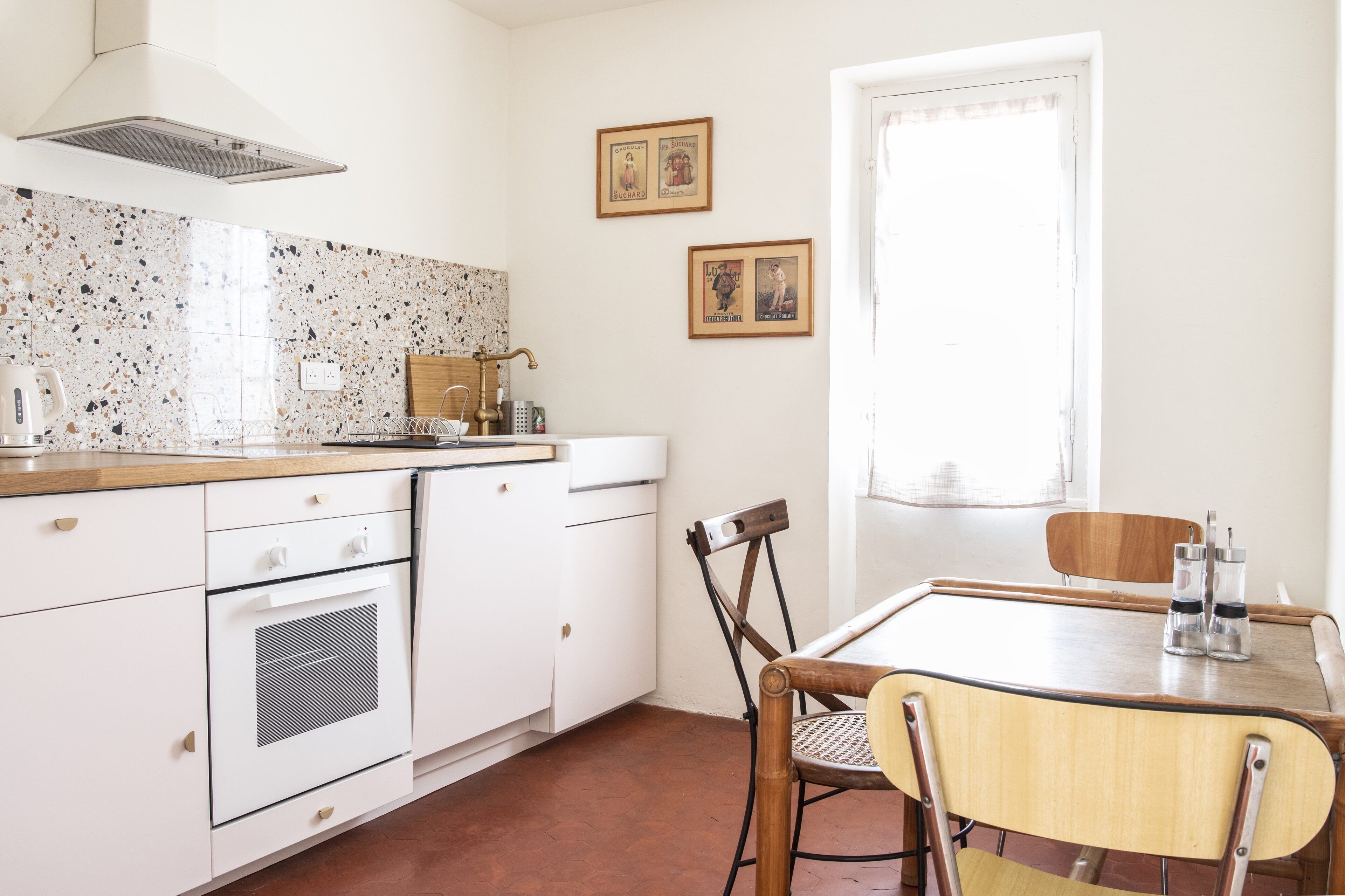 A kitchen with white cabinets, a stove, and a dining table with chairs. There are two framed pictures on the wall and a window with curtains.