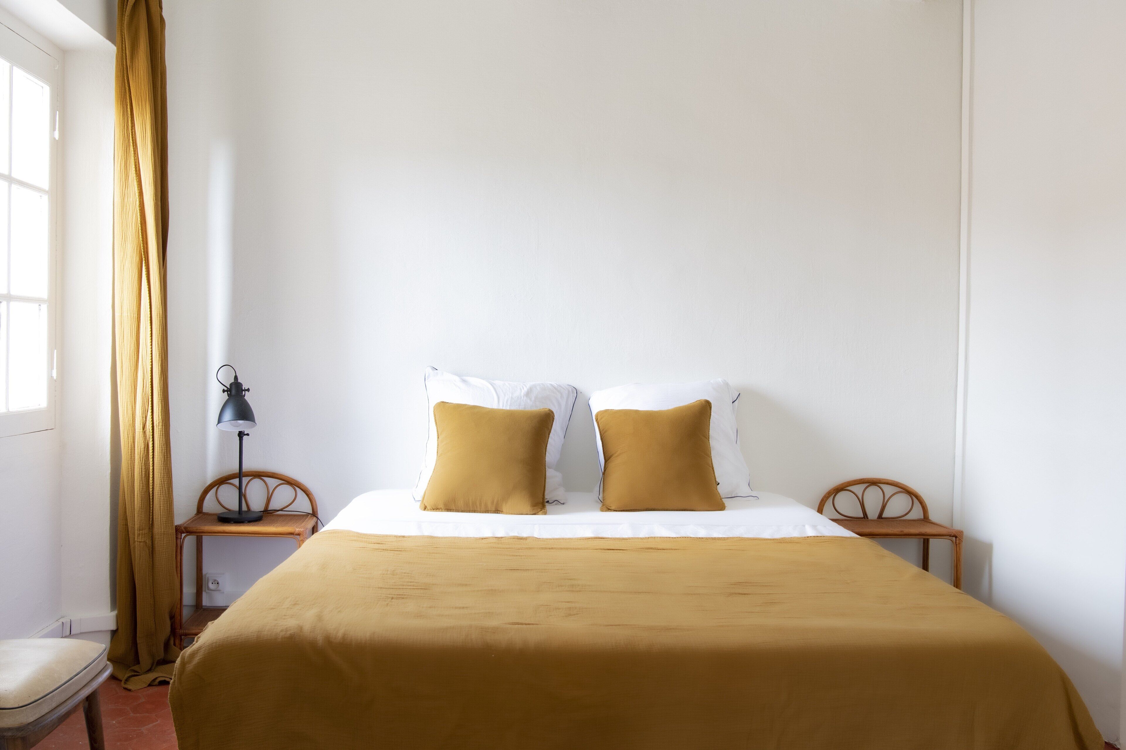 A neatly made bed with a brown bedspread and two pillows, a small wooden nightstand with a lamp, a chair, and a window with curtains.