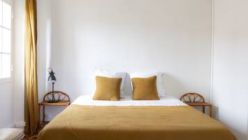 A neatly made bed with a brown bedspread and two pillows, a small wooden nightstand with a lamp, a chair, and a window with curtains.