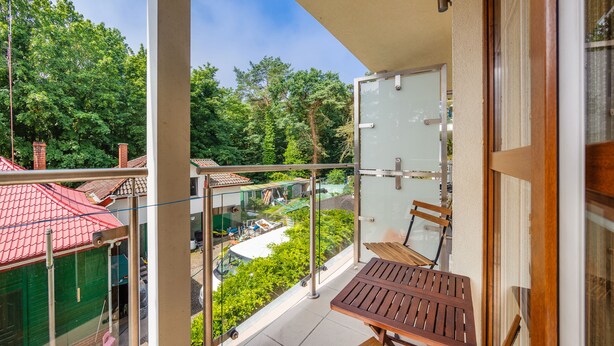 A balcony with a glass door, a wooden table, and a view of a green garden and buildings. - Apartamenty Sun&Snow Rezydencja Bursztyn (Swinoujscie)