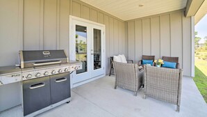 Eine überdachte Terrasse mit Grill aus Edelstahl, Korbmöbeln und einer Schiebetür, die in einen Garten führt.