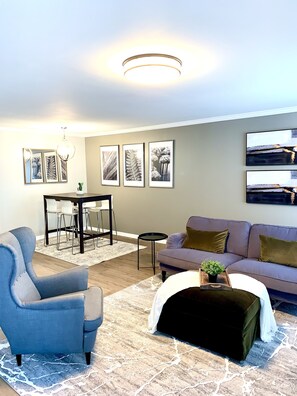 Una sala de estar con sofá, sillón, mesa de centro, mesa de comedor, sillas y cuadros en la pared.