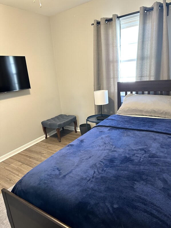 Un dormitorio con cama, televisor montado en la pared, una mesita de noche con lámpara y ventana con cortinas.