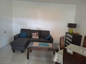 A living room with a dark grey sofa, two blue cushions, a wooden coffee table, and a small cabinet with a lamp.