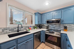 Una cocina con armarios azules, una estufa de acero inoxidable, lavavajillas y una ventana con persianas.