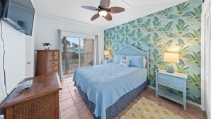 Ein Schlafzimmer mit Bett, Nachttisch, Deckenventilator, Fernseher, einem Holzschrank, einem Fenster und einer Tür, die auf einen Balkon führt.