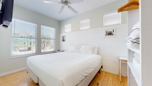 Une chambre à coucher avec un lit, des murs blancs, un plancher en bois, un ventilateur de plafond et plusieurs fenêtres équipées de stores.
