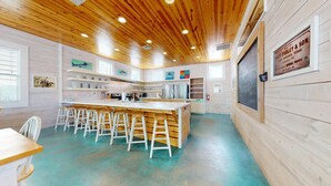 Une cuisine moderne avec un plafond en bois et des murs blancs, comprenant un îlot central avec des tabourets de bar, des appareils en acier inoxydable et des œuvres d’art accrochées aux murs.