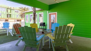 Un espace extérieur aménagé avec une zone de repos couverte, des chaises Adirondack colorées et une table, un petit barbecue, et un bâtiment vert en arrière-plan.