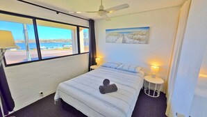 A hotel room with a bed, two bedside lamps, a ceiling fan, a painting on the wall, and a view of the sea through large windows. - Riverview Holiday Waterfront Apt 23 - 1st Floor - Kalbarri WA (KALBARRI)
