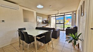 A dining area with a table and chairs, a sofa, a TV, and a view of a playground through a glass door. - Riverview Holiday Apartment 32 - Ground Floor -  Kalbarri WA (KALBARRI)