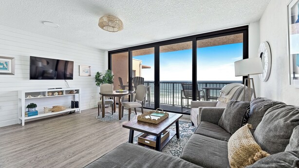 A modern living room with a large window, a flat-screen TV, a sofa, a coffee table, and a dining area with chairs. - 🏖️Oceanfront Sunrise Overlooking Pool!  Car Free Beach, Walk to shopping & dining! Sunrise 805 (New Smyrna Beach)