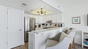 A modern kitchen with white cabinetry, a stainless steel refrigerator, and a central island with bar stools. - 🏖️Oceanfront Sunrise Overlooking Pool!  Car Free Beach, Walk to shopping & dining! Sunrise 805 (New Smyrna Beach)