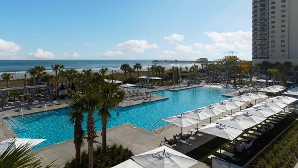 A resort pool area with multiple lounge chairs and umbrellas, a clear view of the ocean, and a tall building in the background. - Re-decorated Ocean View Unit-Full Access-605B (Myrtle Beach)