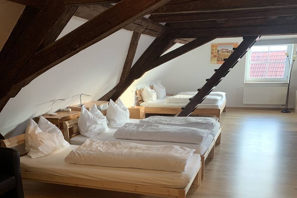 Quarto com piso de madeira, teto inclinado e várias camas dispostas em fila.