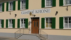 A building with a sign reading "Gasthof KRONE" and a wooden door with a metal staircase leading to the entrance.