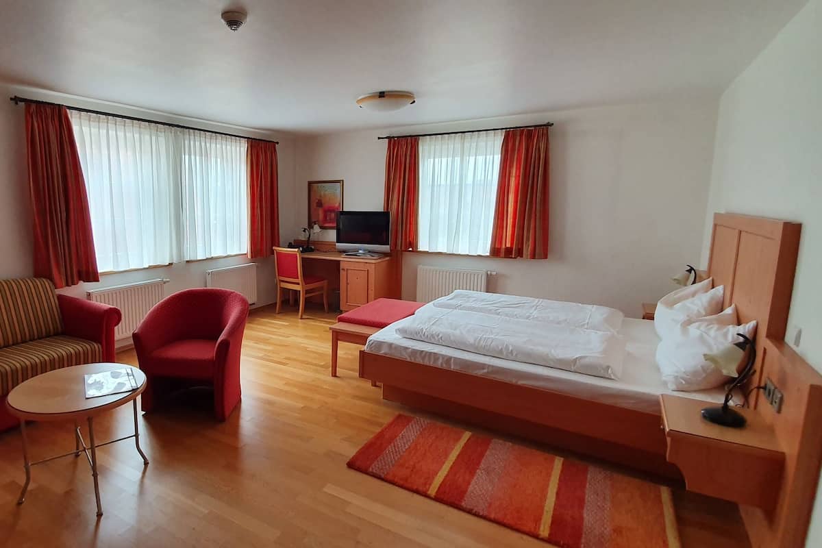 Quarto de hotel com cama, sofá, poltrona, escrivaninha com computador e janela com cortinas.