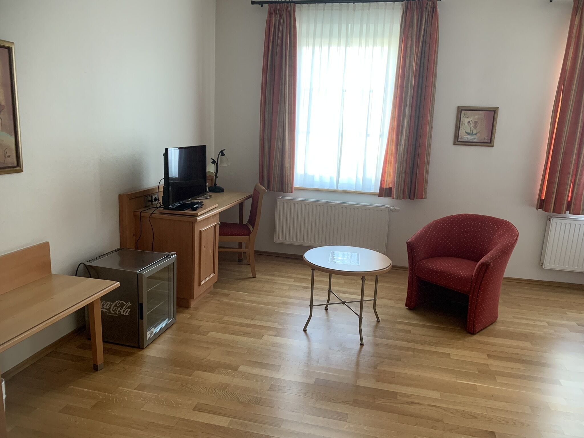 Una habitación amoblada con un escritorio de madera, un televisor, un sillón rojo, una mesita y un radiador.