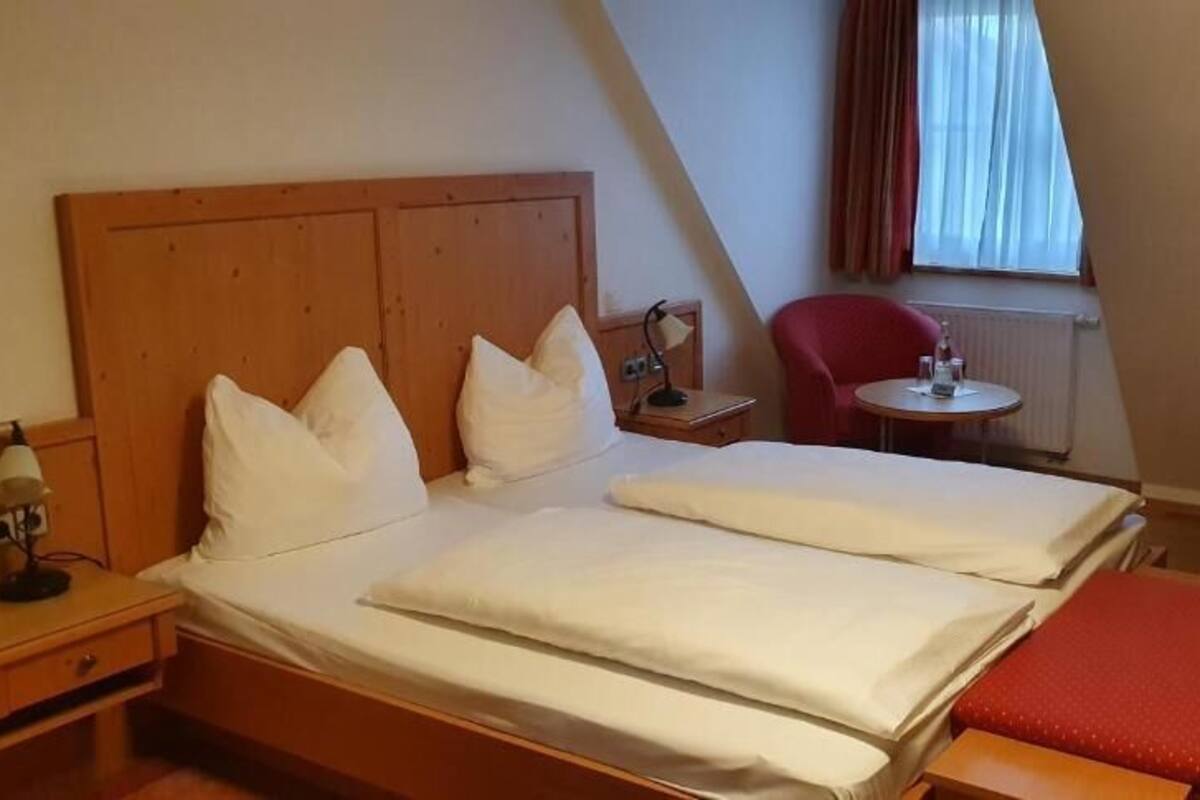 Quarto de hotel com cama de madeira, uma cadeira vermelha, uma mesinha e uma luminária.
