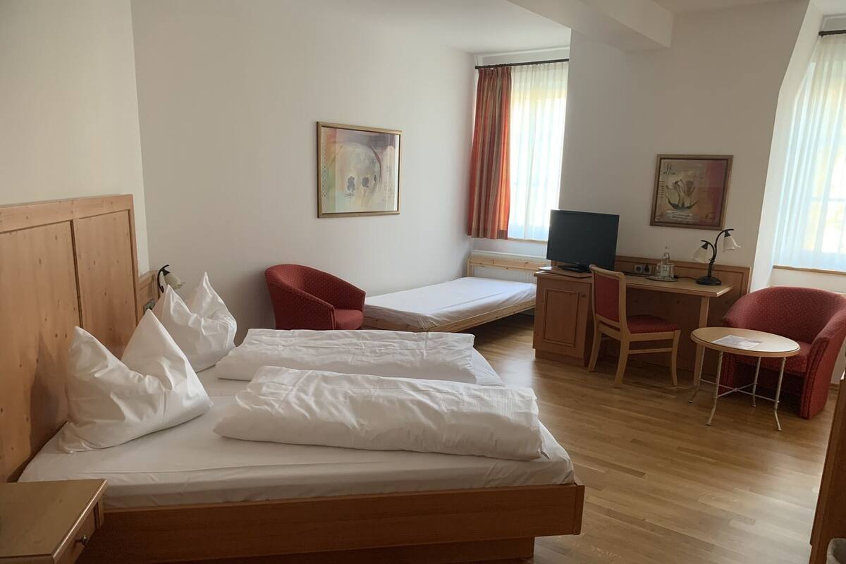 Quarto de hotel com cama de madeira, uma escrivaninha com computador, uma cadeira e uma pequena mesa.