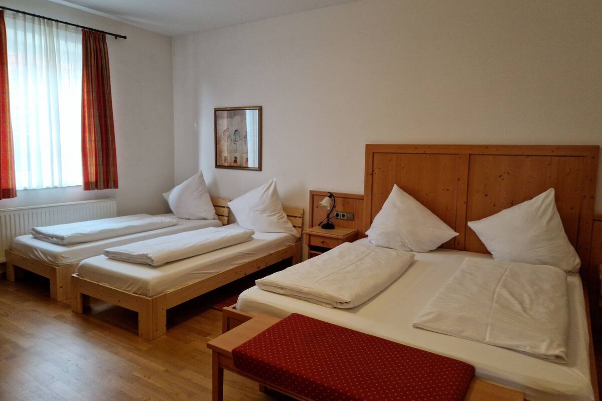 Quarto de hotel com duas camas de solteiro e uma cama de casal, cabeceiras de madeira, uma pequena mesa de cabeceira com abajur e uma janela com cortinas.