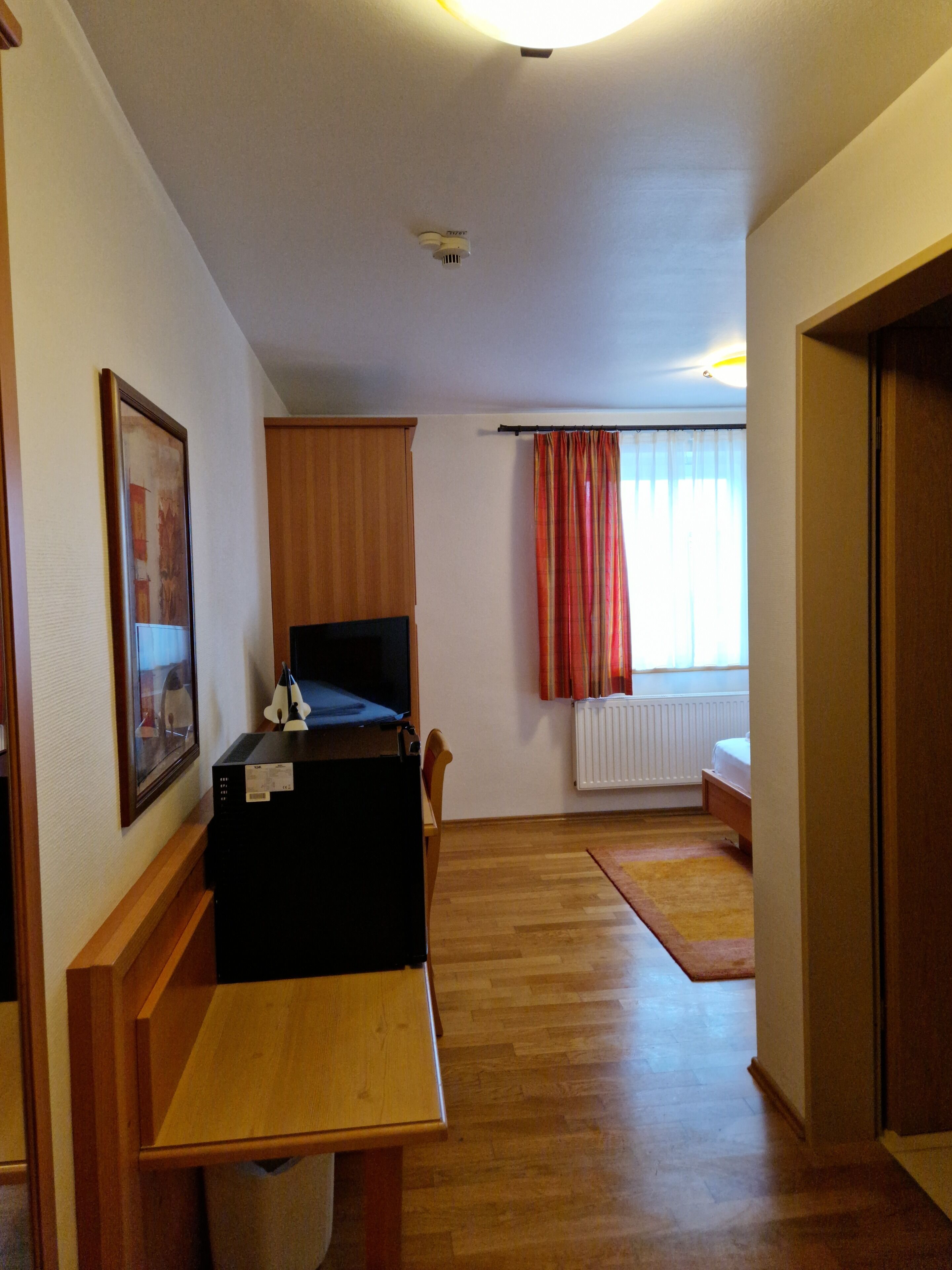 A room with a wooden desk, a television, a radiator, and a window with curtains.