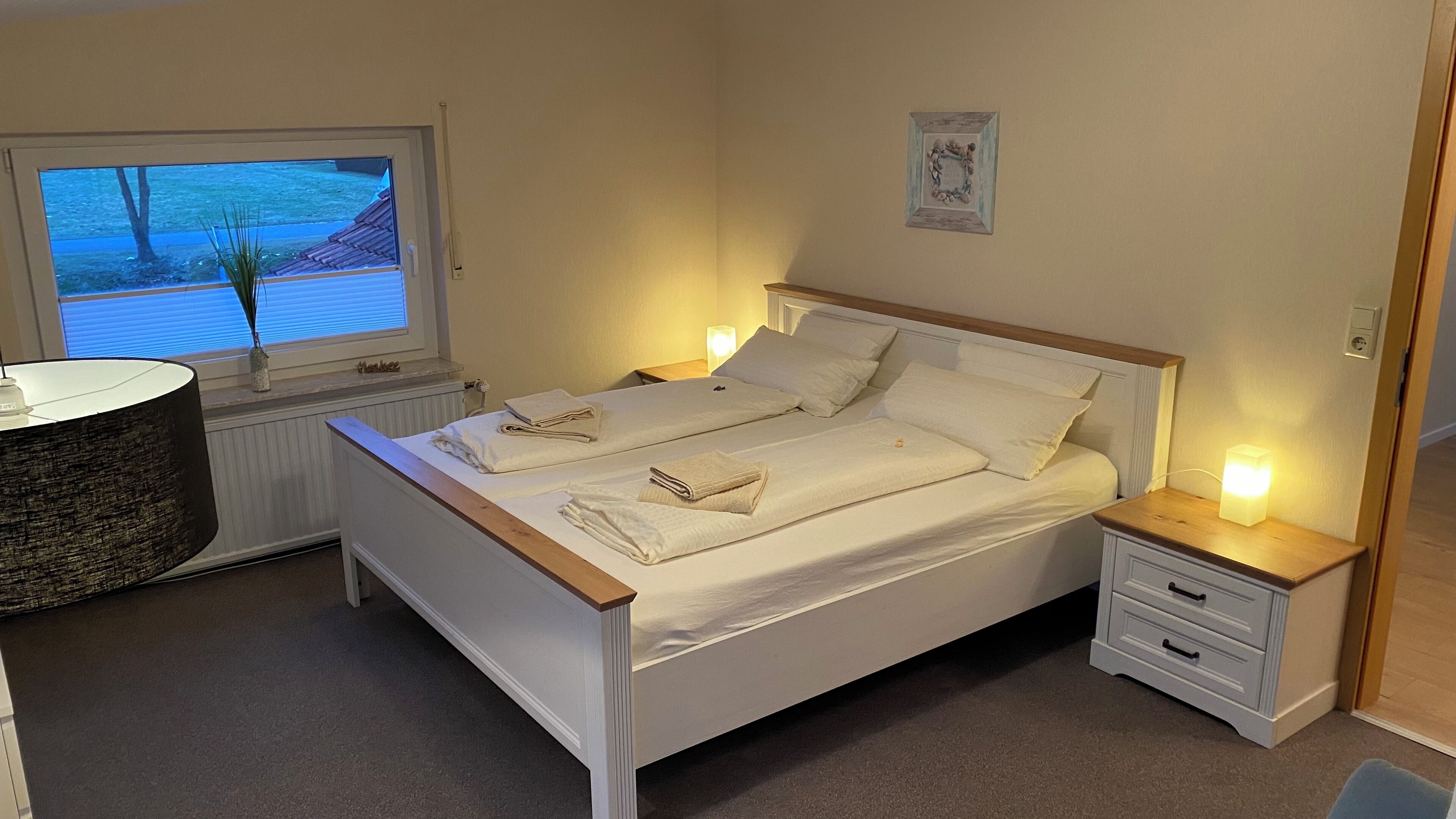 Un lit double avec du linge de lit blanc, deux tables de chevet avec des lampes, une fenêtre avec des stores et un radiateur.