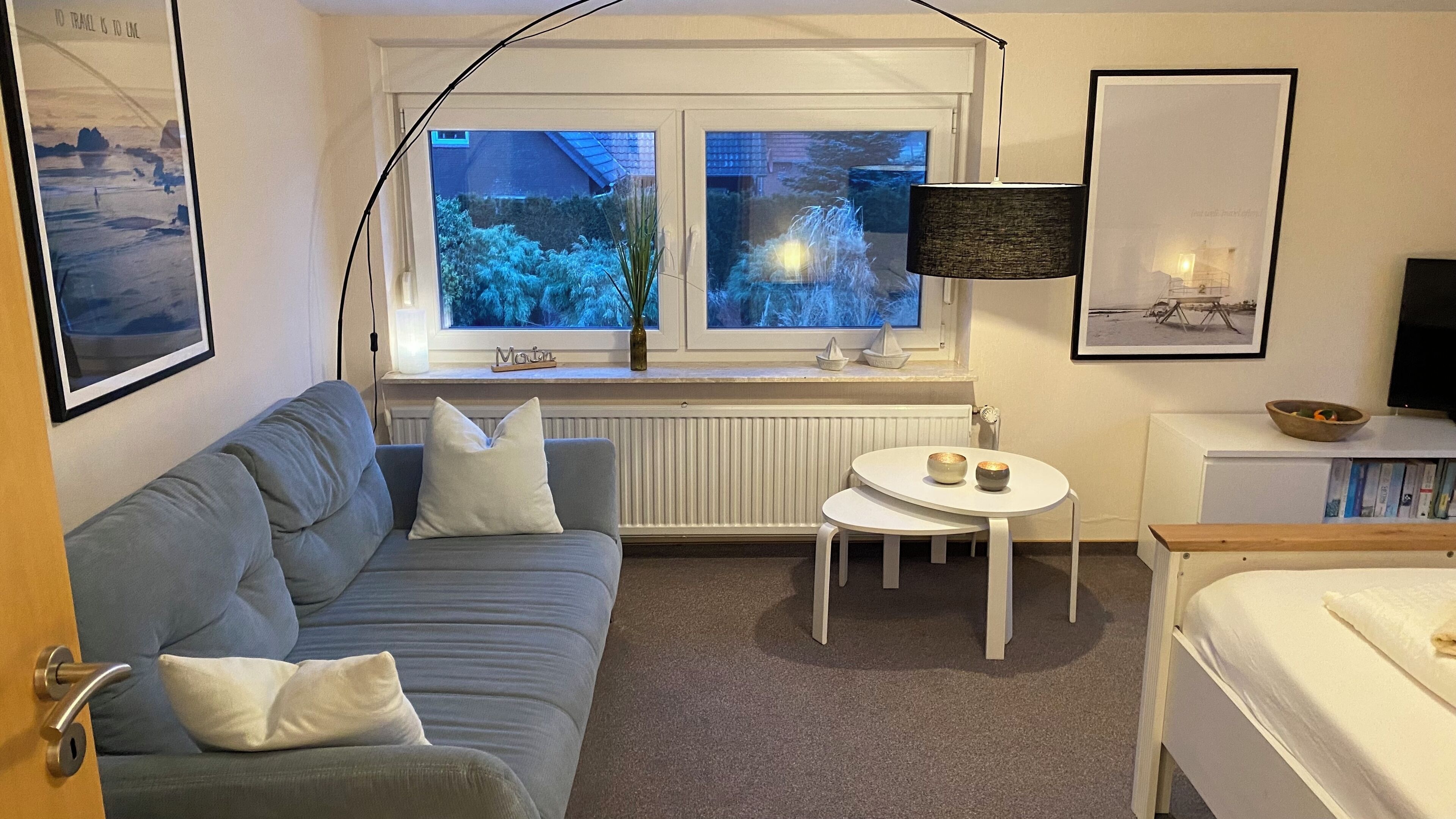 Une pièce chaleureuse avec un canapé gris, une petite table ronde sur laquelle sont posés deux bols, un radiateur blanc et une fenêtre donnant sur des arbres.