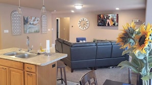 A kitchen with a double sink, wooden cabinets, and a built-in island. There is a dark-colored sofa in the background and a large clock on the wall.