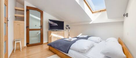 Un dormitorio con tragaluz, una cama con sábanas blancas, un televisor, un ventilador de pie y un armario de madera.