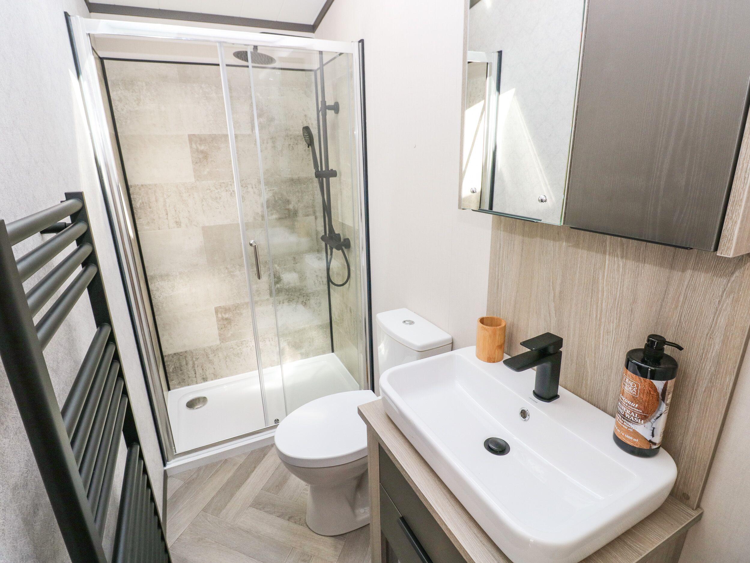 A modern bathroom with a glass shower enclosure, a white toilet, a sink with a black faucet, and a wooden cup holder.