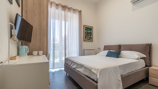 Een slaapkamer met een bed, een bureau met een computer monitor, een koffiezetapparaat en een schilderij aan de muur.