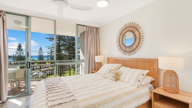 A hotel room with a large bed, two bedside lamps, a decorative mirror, and a view of the ocean through a sliding glass door.