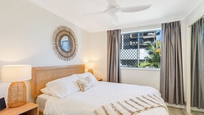 A hotel room with a bed, two bedside lamps, a ceiling fan, a window with curtains, and a decorative mirror on the wall.