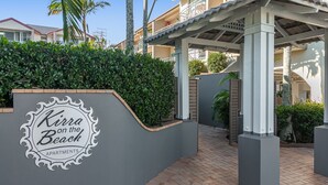 A sign for 'Kura on the Beach Apartments' with a modern architectural design, including a covered walkway and a landscaped area with palm trees.