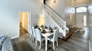 Un vaste espace dédié à la salle à manger, avec une table et des chaises blanches, un escalier et une porte donnant sur une autre pièce.