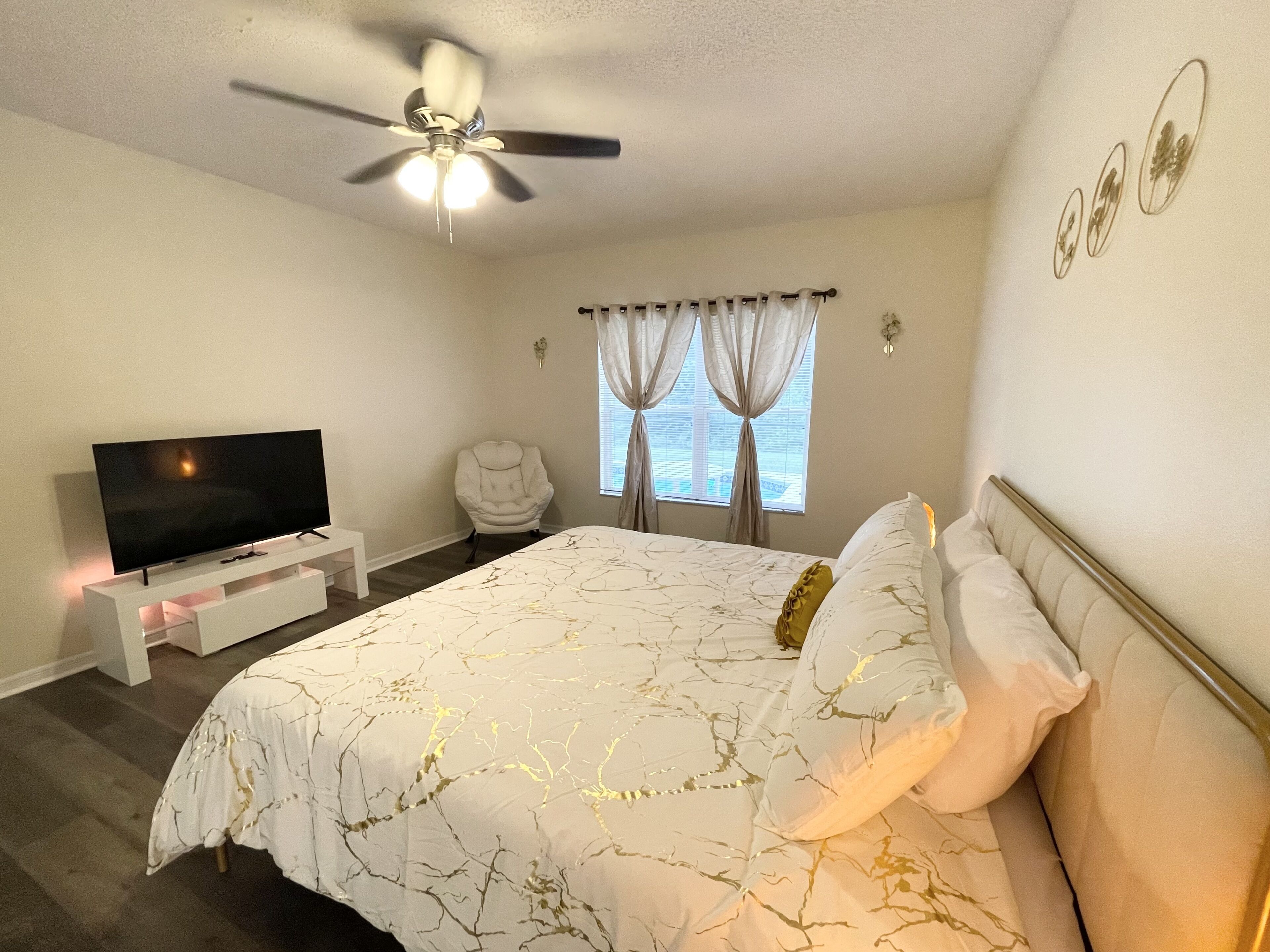 A bedroom with a bed, a TV on a stand, a ceiling fan, and a window with curtains.