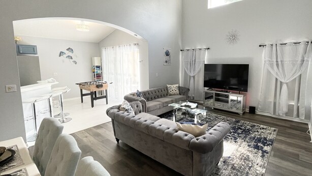 Un salon spacieux avec un canapé gris, une table basse et un téléviseur à écran plat. On y trouve également un coin repas avec un bar et des chaises, ainsi qu’une grande fenêtre qui laisse entrer la lumière naturelle.