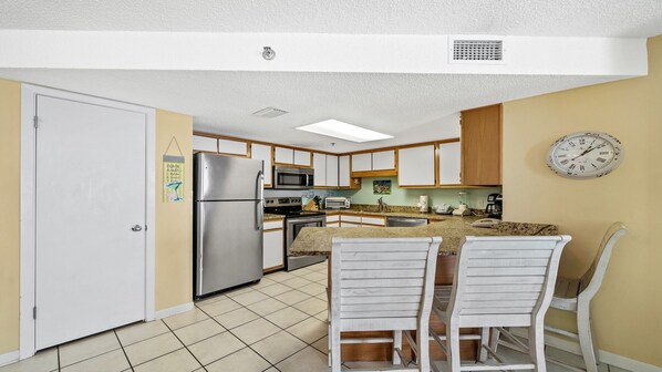 A compact kitchen with a refrigerator, microwave, and stove. There are two white chairs and a small dining table. - Snowbird Availability! Beachfront 3BR!! Splash Pad (Orange Beach)