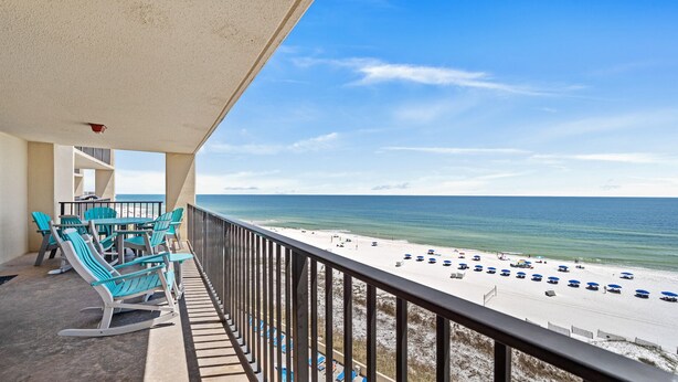 Un balcon avec des chaises turquoise et une table donnant sur une plage parsemée de parasols et de chaises longues.