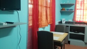 Ein Zimmer mit an der Wand befestigtem Fernseher, einem Esstisch mit Stühlen, einer Küchenzeile mit Regalen und einem Fenster mit roten Vorhängen.